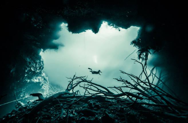 Entrenamiento de buceo técnico en cuevas: buzo revisando manómetros en configuración de doble tanque (sidemount) en entorno bajo techo.